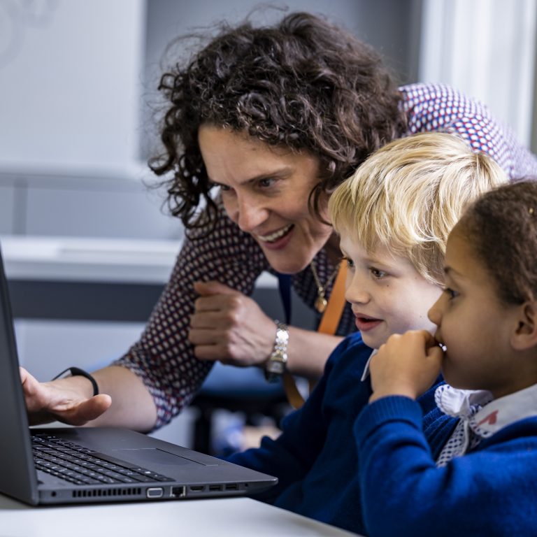 2 children using a laptop, with a teacher helping out